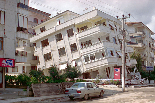 A white, four storey building in Izmit, Turkey, leans at a dangerous angle due to an earthquake. There is a road next to it with other buildings that are not affected.