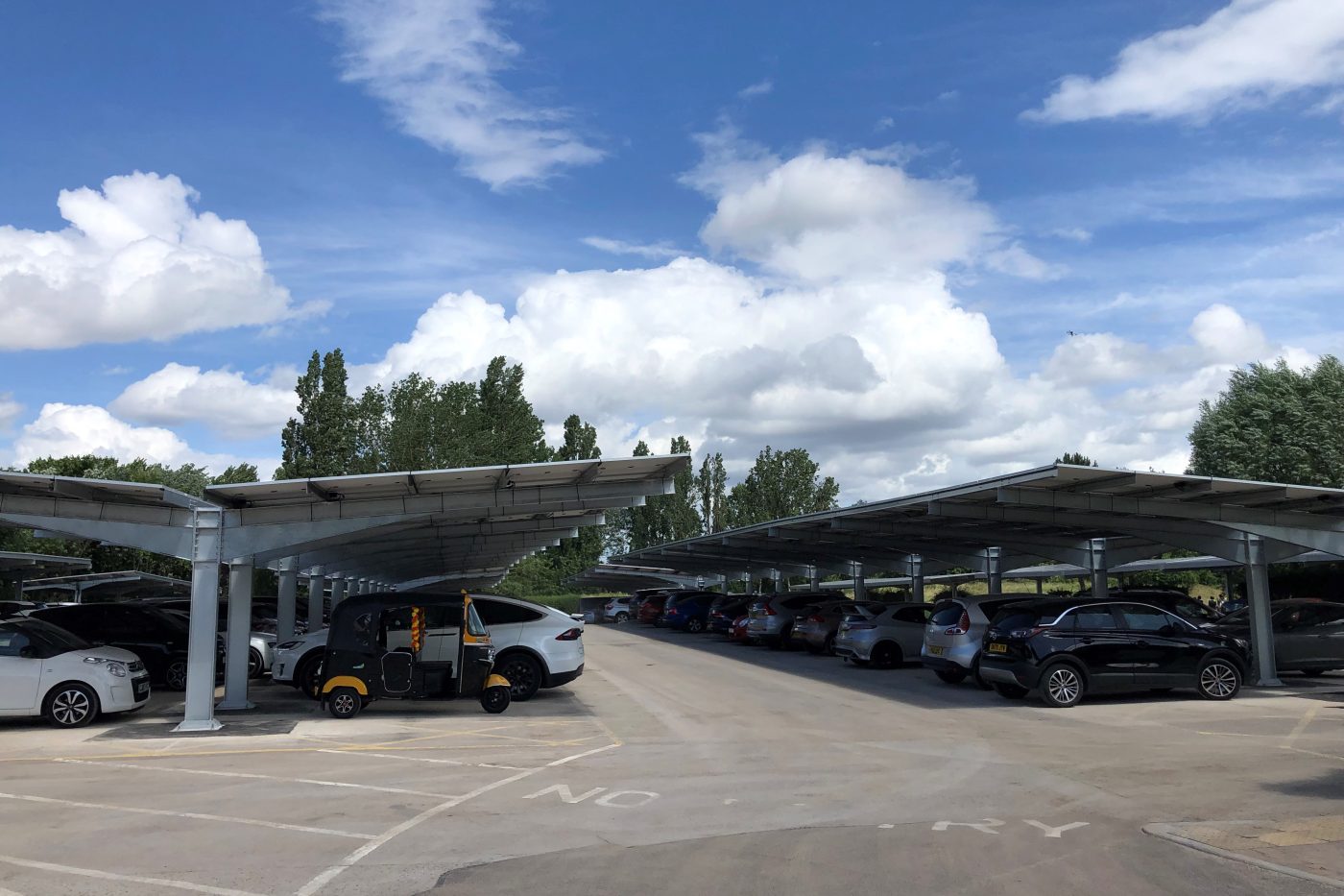 Cars parked in a car park beneath open-sided structures (car ports) with roofs covered in solar panels. The sky is bright, with big fluffy clouds.